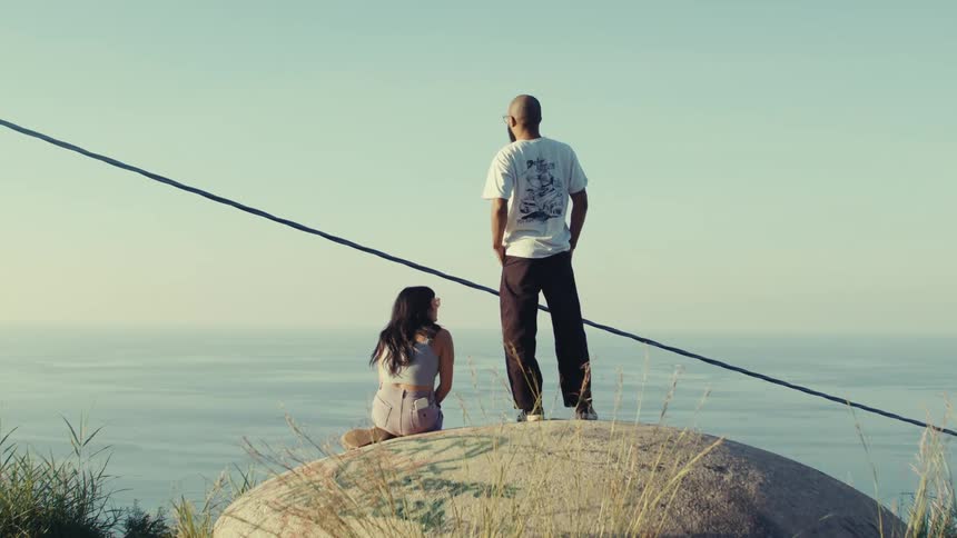 A couple is standing on a large rock overlooking a vast calm ocean under clear skies