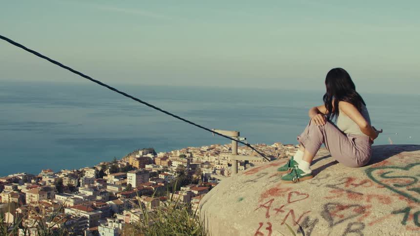 A woman is sitting on a rock with graffiti overlooking a coastal town with buildings and the sea in the background