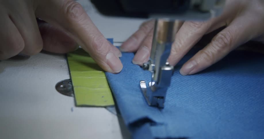 A persons hands guiding blue fabric under a sewing machine needle while trimming excess material with a small tool