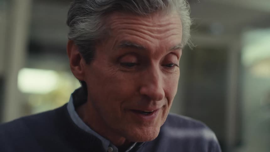 A man with gray hair wearing a dark jacket is standing outdoors having a conversation