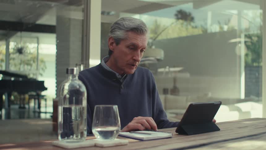An older man with gray hair sits at an outdoor table using a tablet engaged in a video call