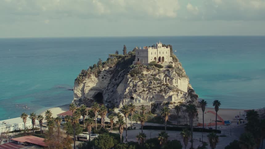 A drone shot reveals a coastal scene panning down from a mostly clear sky to a building perched atop a rocky outcrop overlooking the sea