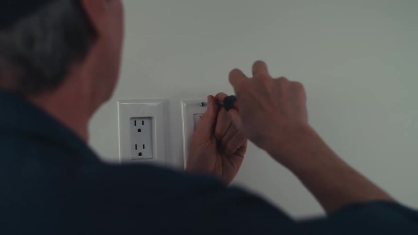 A man in a cap stands with his back turned and uses a screwdriver to tighten a screw in a wall