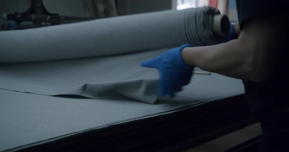 A person wearing blue gloves is unrolling a large piece of gray fabric on a table