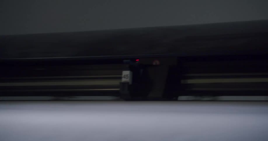 A cutting or printing plotter with a red light is running above a sheet of white material