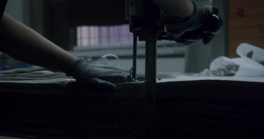 A gloved hand uses an industrial fabric cutter to cut cloth layers on a large table