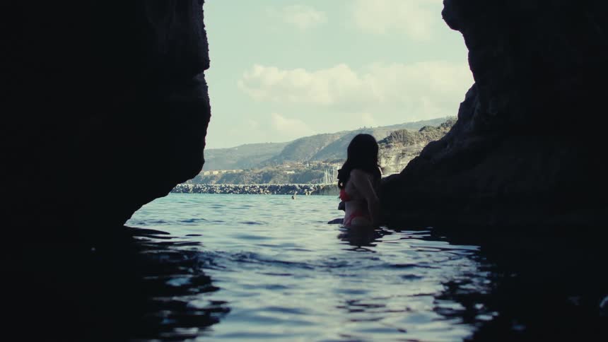 A woman in a bikini stands hip deep in water inside the entrance to a cave