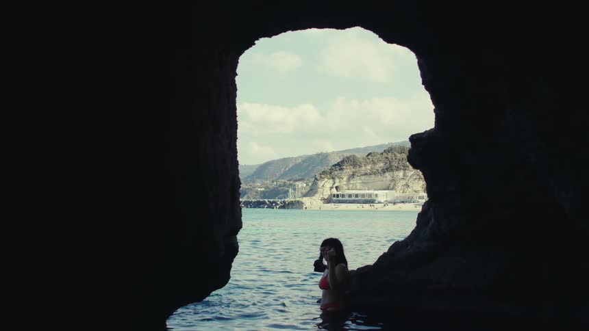 A woman in a bathing suit stands in the water near the exit of a dark stone cave