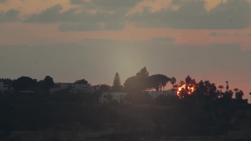 A wide shot captures a sunset behind a hilltop silhouetting houses and trees