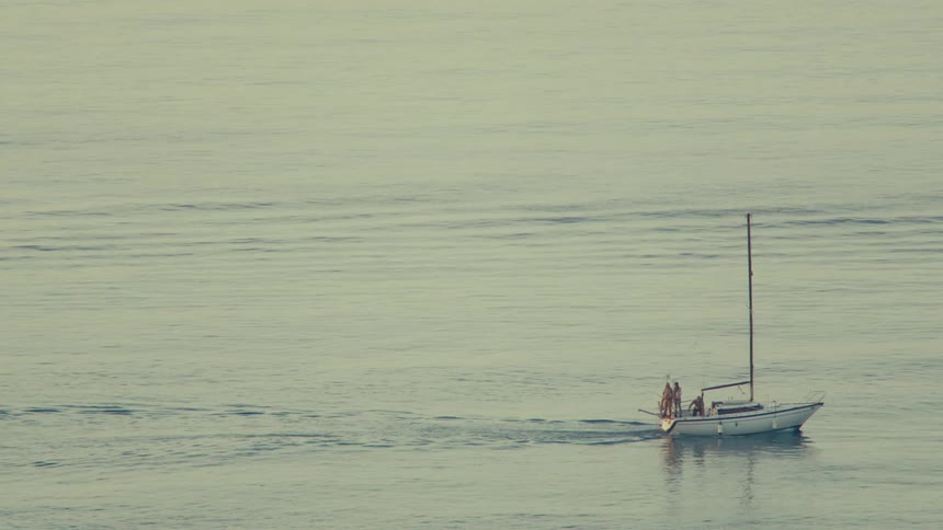 A small white boat with the sail furled and some people on top of it and it is sailing on a sea with calm water