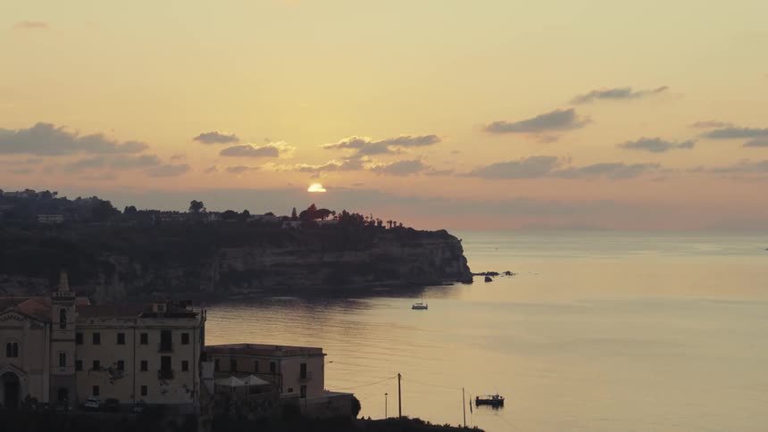 Sunset over a seaside cliff with an old building in the foreground and some boats in the calm water