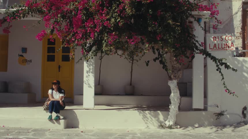 Woman wearing a cap and sunglasses sitting on the porch of a white house next to pink flowers and a tree outdoors during daytime