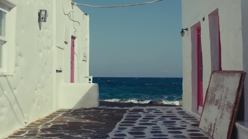 A narrow alleyway between two white buildings with pink doors leading to a view of the ocean with waves crashing against the shore