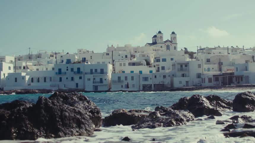 Sea water splashes over shore side rocks near a coastal town on a slightly cloudy day