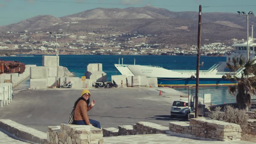 A woman in a yellow hat sits on a low stone wall looking out at a ferry docked at a harbor with a town and mountains in the distance