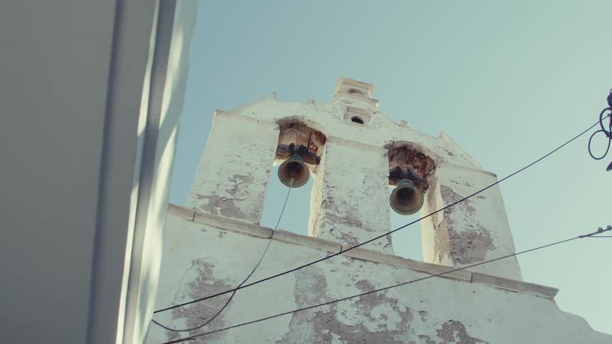 N upward view of a weathered whitewashed bell tower with two bells set against a clear blue sky with electrical wires crossing in front of it