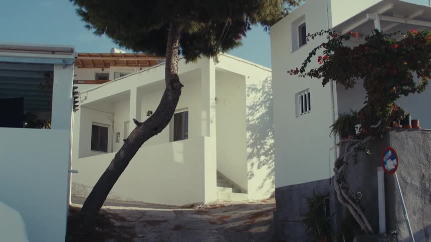 A whitewashed building with a redroofed house in the background and slanted trees in the front