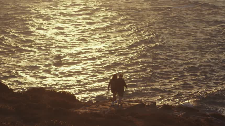 Two people walking toward the coast are partially silhouetted against sunlight reflecting ocean water