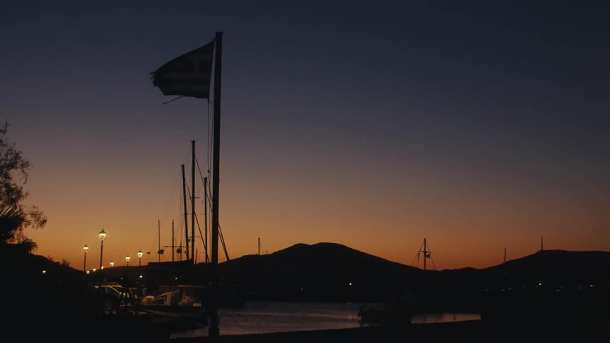Flag flutters in the wind at the dock located near hills silhouetted against a red sky at night