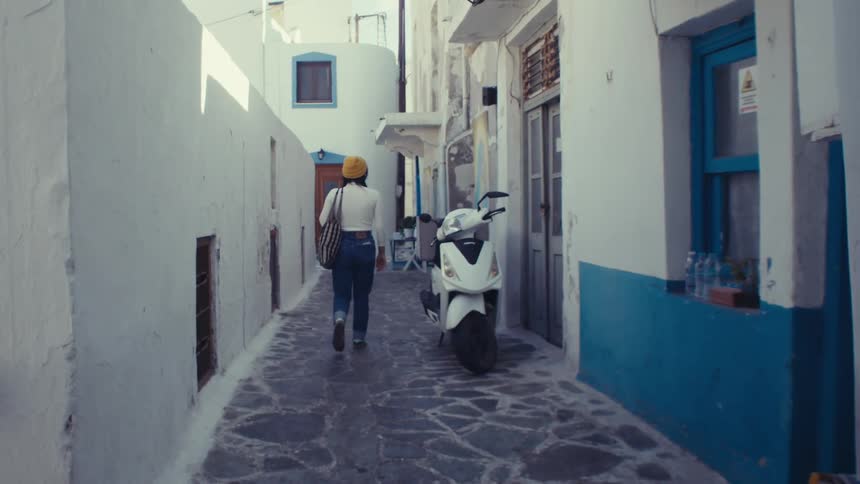 A woman in a yellow hat a white shirt and jeans walks down a narrow whitewashed street in a greek village passing a parked scooter