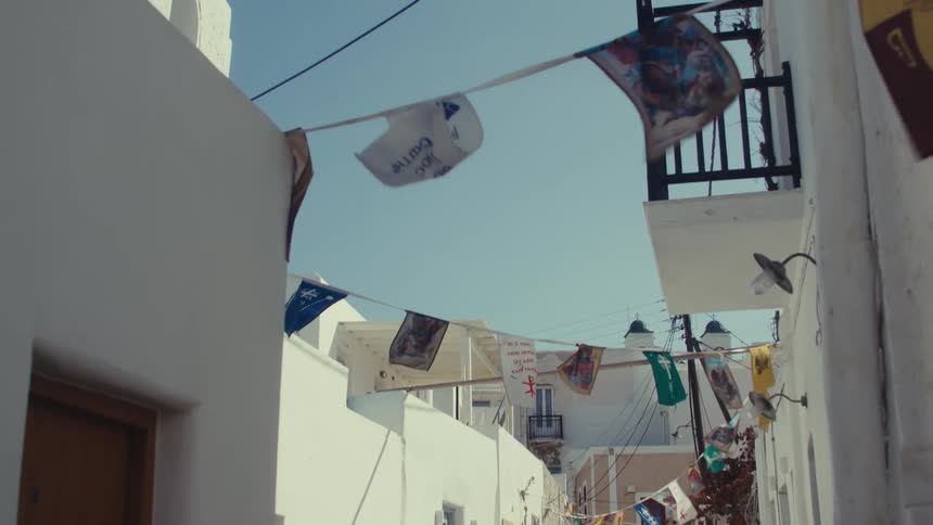 A narrow sunlit alleyway in a mediterranean village is adorned with colorful flags and lined with whitewashed buildings