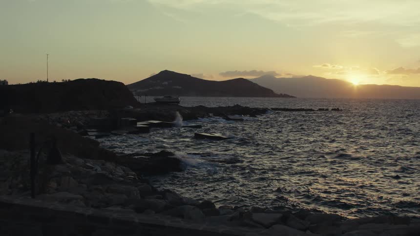 A picturesque sunset casts a golden glow over a rocky coastline with a distant mountain range silhouetted against the sky