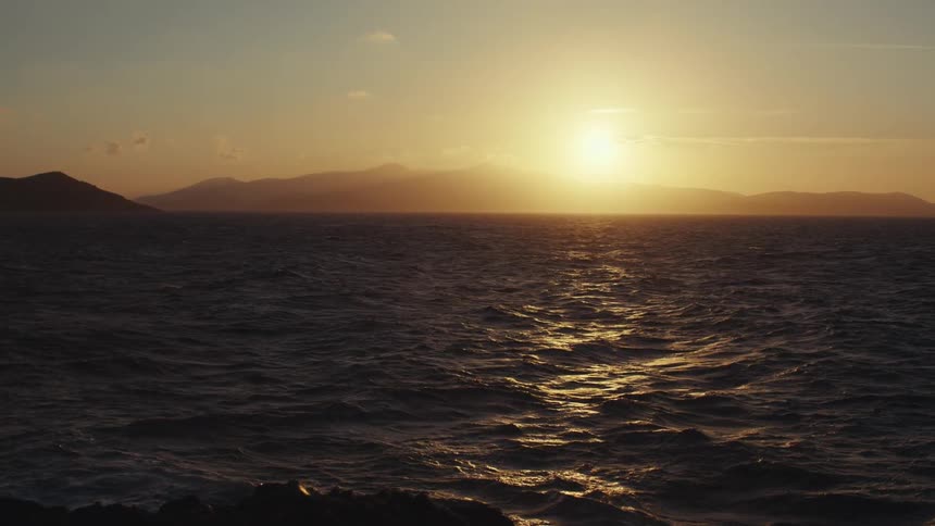 Sun hovers over seaside mountains in a red tinted sky and shines onto ocean waves