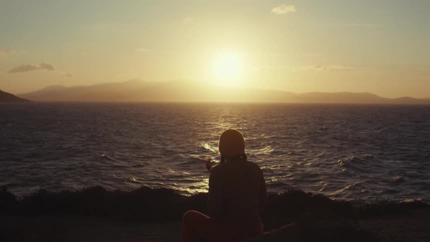 A person sits silhouetted against a sunset over a calm sea and distant mountains