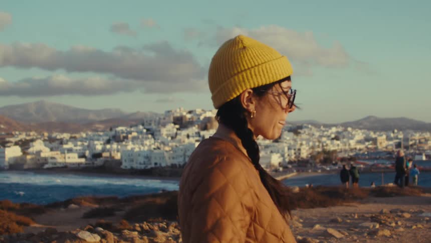A woman wearing a yellow beanie and glasses stands on a hill overlooking a whitewashed coastal town and the sea