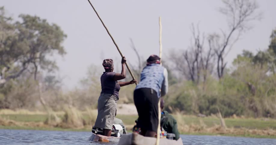 Two boats with people standing using huge sticks to row while there are some people sitting in the boats