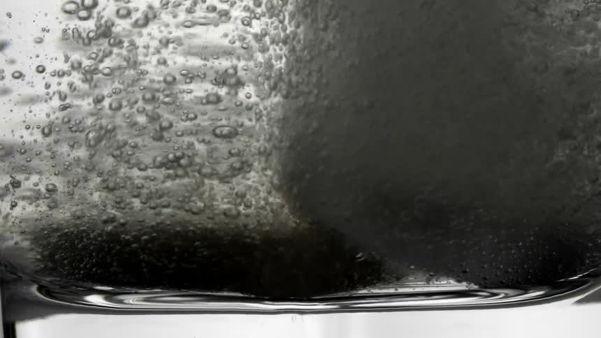 A dark colored tablet dissolves in a glass of water releasing numerous bubbles