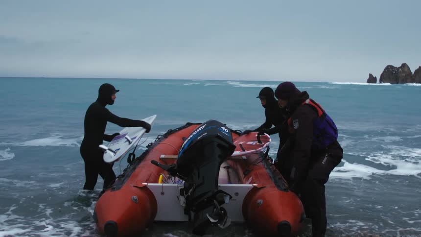 Three individuals in wetsuits are preparing an orange inflatable boat with an outboard motor on a rocky shore
