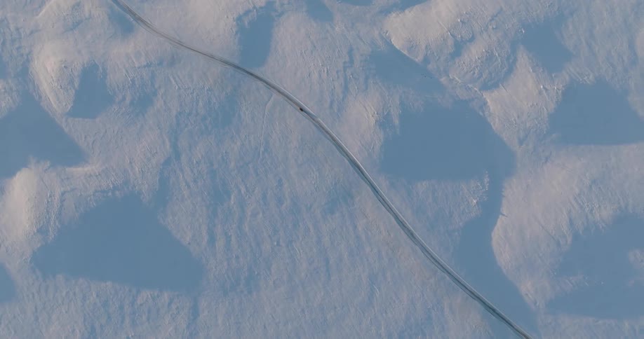 An aerial view of a large asphalt road in the middle of a completely snowy region where there is a car driving along it