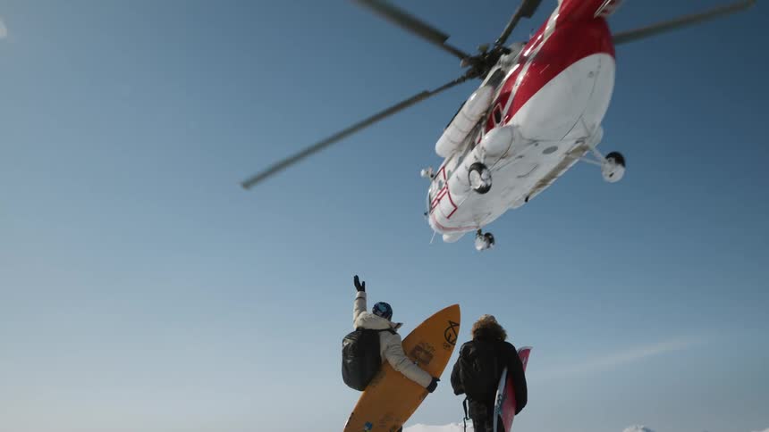 Two individuals with surfboards and backpacks stand on a snowy mountain looking up at a helicopter flying overhead