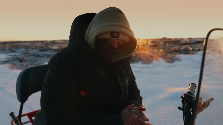 A person is sitting on a snowmobile in a snowy landscape at sunset putting on gloves