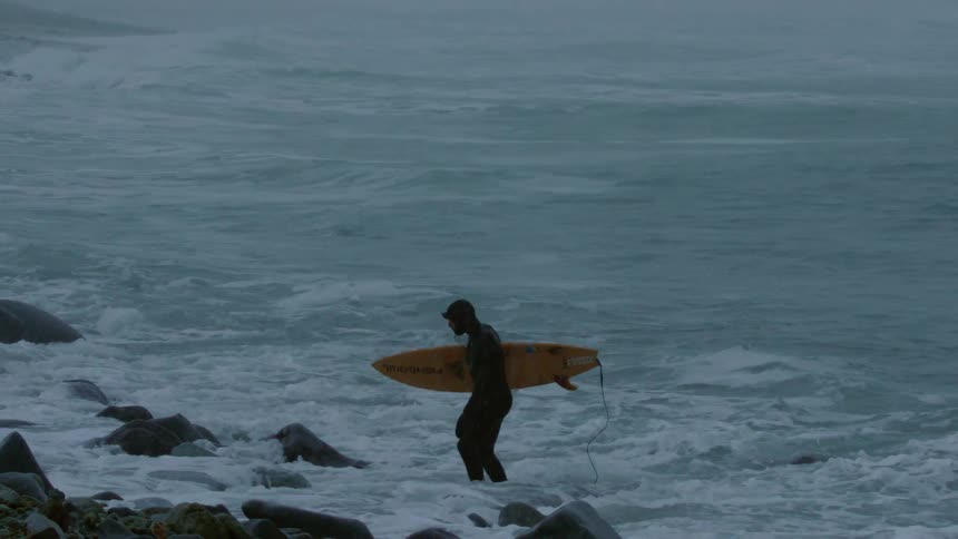 A solitary surfer walking through rocky shallow water on a cloudy day holding a yellow surfboard
