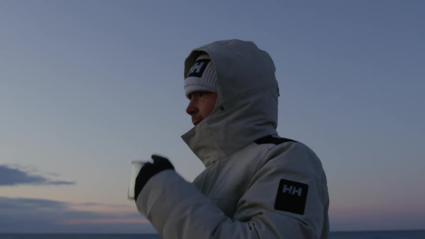 Man wearing a winter jacket sips from a mug while looking toward the red tinted horizon