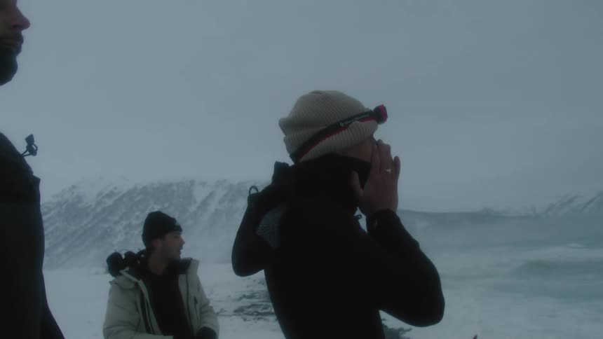 Three men in cold weather gear stand on a snowy beach preparing to enter icy water with mountains visible in the background