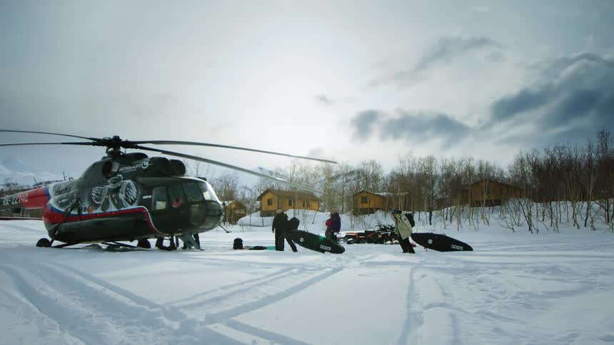 A helicopter parked in a snowy field with people unloading equipment