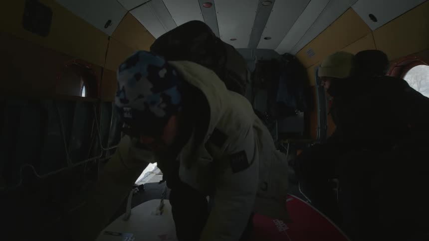 People are stepping into an aircraft from a snowy landscape while carrying two red surfboards and wearing winter gear