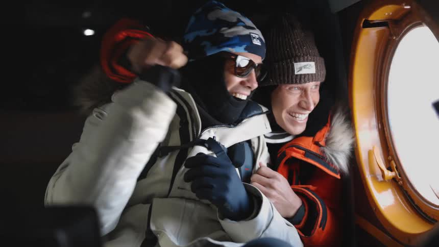 Two men in warm clothing are sitting and smiling as they look out a window