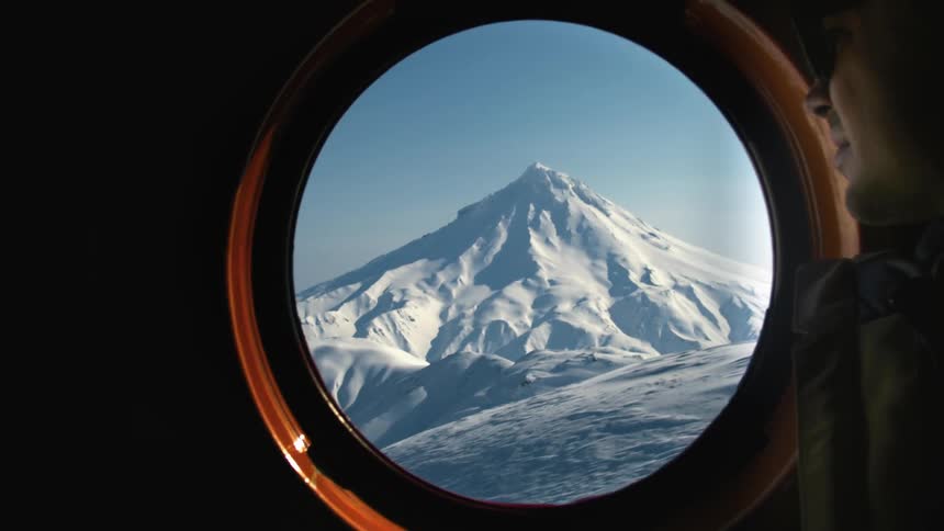 A view of a huge mountain covered in white snow seen through a round window where there is a person watching on the right side