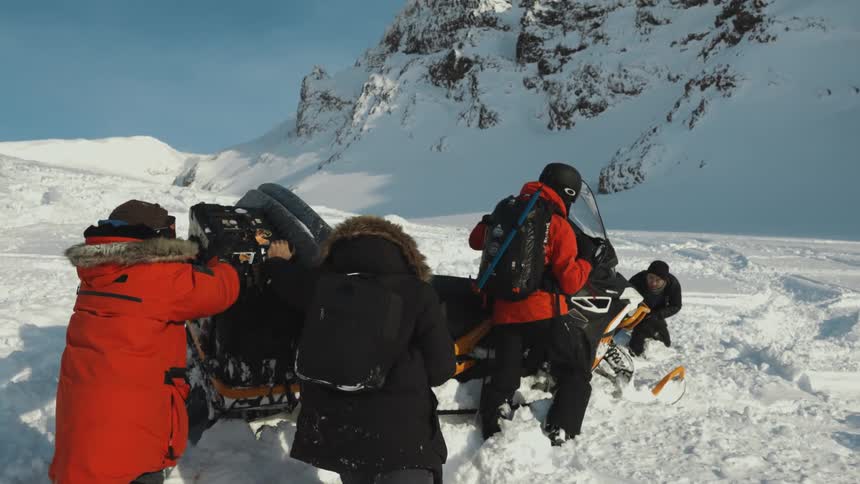 People in winter clothing are pushing a snowmobile that appears to be stuck in deep snow on a mountainous snowy terrain