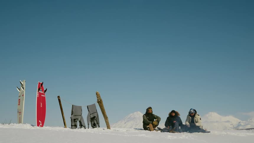 Three people sit around a small fire in a snow covered area next to two snowboards