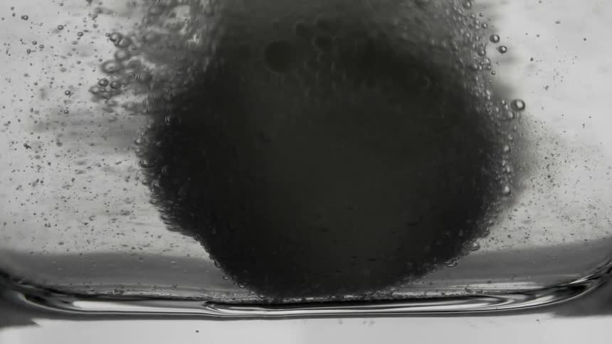 A dark spherical object is submerged in a clear liquid creating ripples and bubbles on the surface