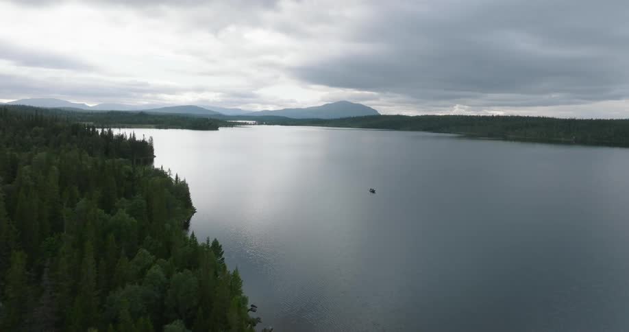 A serene lake is surrounded by forested hills and mountains under a cloudy sky with two people in a small boat on the water