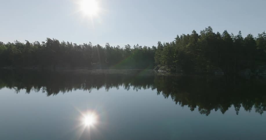 A serene lake reflects the surrounding dense forest and the bright sun in a clear sky