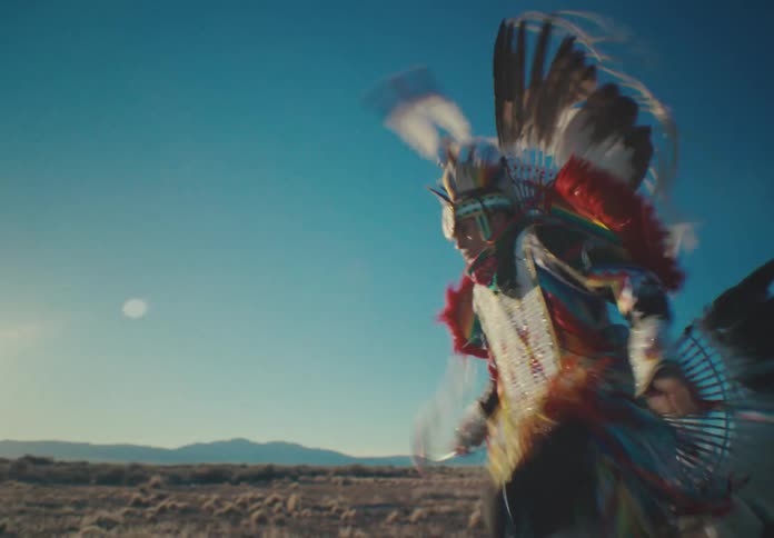 A person in elaborate native american regalia dances in a wide open landscape as the sun sets behind them