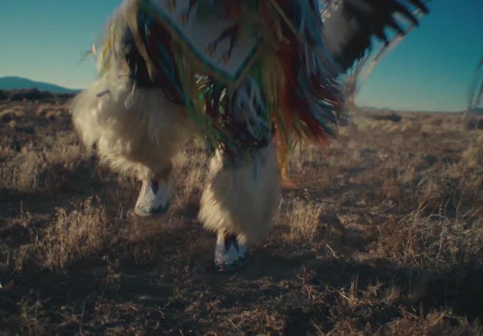 A person is dressed in traditional colorful native american regalia with feathers and intricate patterns dancing in an open field