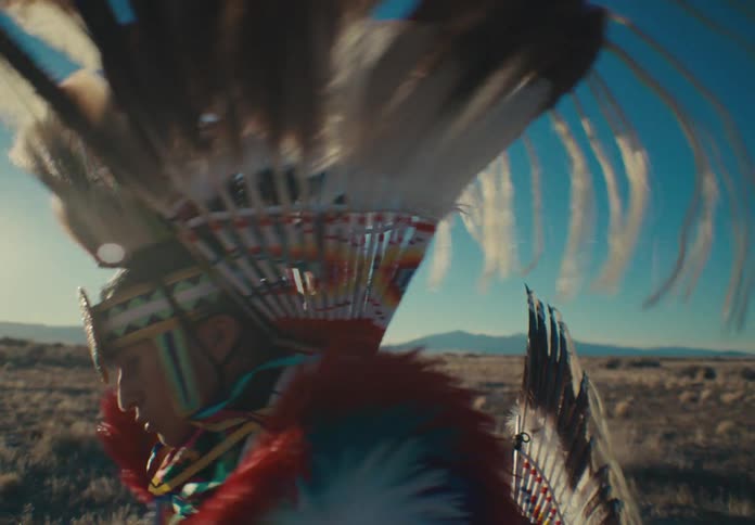 Man wearing traditional native american attire performs outside in a field on a windy day with the mountains on the horizon